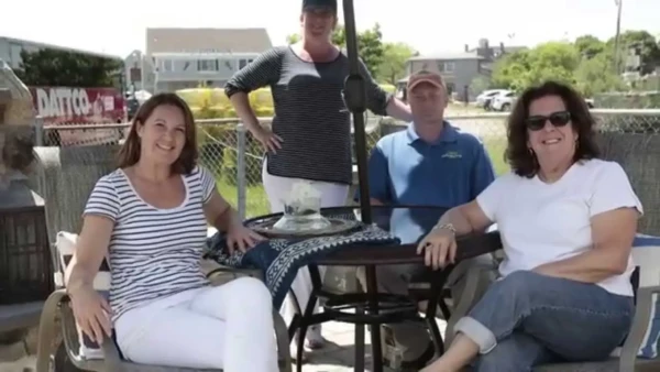 Four people are gathered around an outdoor table under an umbrella, embodying the essence of summer living. Two women sit, smiling, while a woman and a man stand behind them. The sunny setting features a house and lush greenery in the background, making it feel like the perfect event video moment.