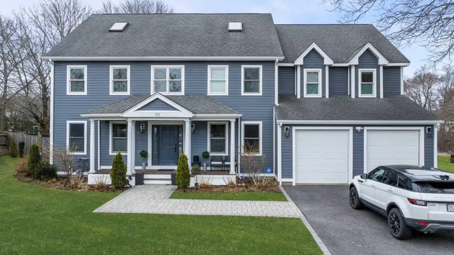 A two-story blue house with white trim, featuring a front porch and two-car garage. A white SUV is parked in the driveway, next to a paved walkway leading to the front door. The lawn is neatly manicured, with bare trees in the background.