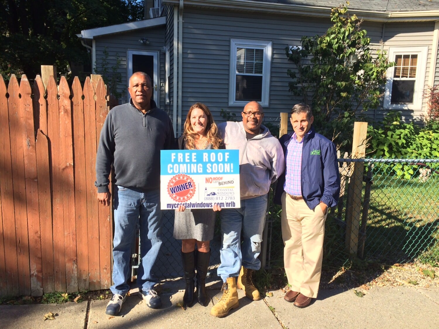 Four people stand outside in sunlight, smiling, holding a sign that says Free Roof Coming Soon! in front of a gray house with a brown fence and blue chain-link gate, celebrating the No Roof Left Behind win from Coastal Windows & Exteriors.