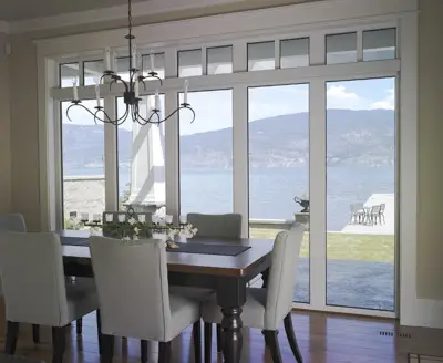 A dining room with a dark wood table, six light gray upholstered chairs, and a modern chandelier. Large high-quality windows offer a scenic view of a lake, mountains, and an outdoor patio with a table and chairs.