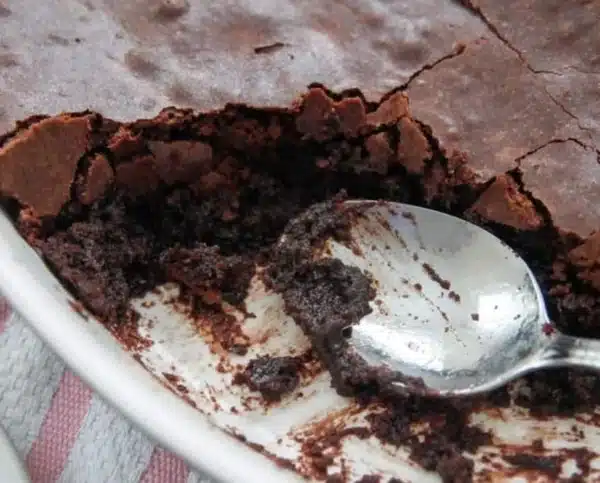 A close-up of a partially eaten Depression Era Chocolate Cake in a white dish, with a spoon resting inside, reveals a gooey, rich texture beneath its cracked top layer.