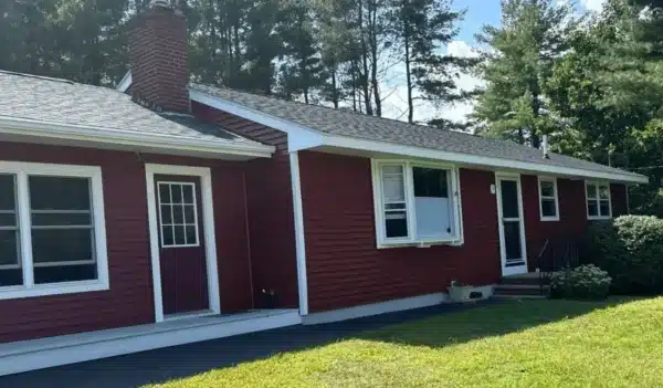 A single-story red house with white trim, black roof, and a brick chimney sits on a sunny day. Featuring replacement bay windows often seen in New England homes, it has a small porch with steps, a green lawn, and tall trees in the background.
