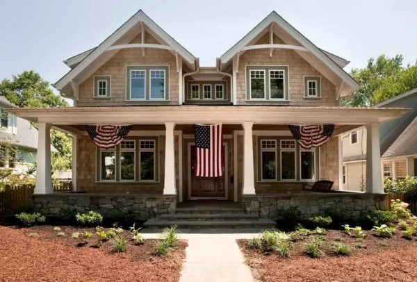 A two-story house with shingle siding, stone foundation, and a wide front porch decorated with three American flags. Fresh landscaping and a concrete walkway lead to the wooden front door—perfect for showcasing top window contractor reviews.