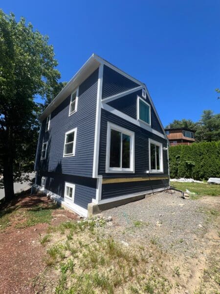 A blue, two-story house with white trim sits under a clear blue sky. The house has large windows and a gravel area at the front. There are trees to the left and a wooden fence to the right.