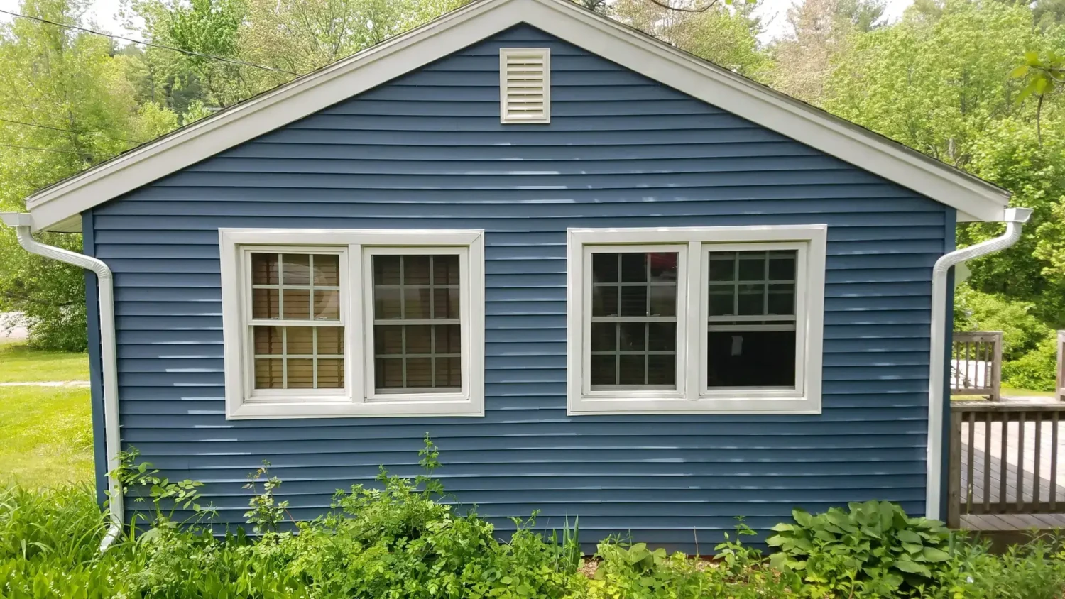 The Best Replacement Windows Buying Guide for New England Homes A blue house with white trim features two double-pane windows and a small vent at the top. Green plants and grass grow in front, while trees and shrubs provide a lush backdrop—ideal for those referencing the best replacement windows buying guide.