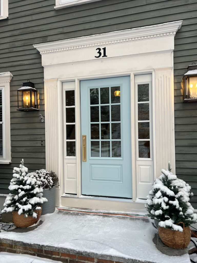 A pastel blue front door with glass panels on a green house, flanked by two small, snow-covered evergreen trees in baskets. The house number 31 is displayed above the door. Two lantern-style lights are on either side. Snow is on the ground.