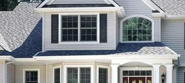 Front view of a two-story house with gray siding and a prominent gable, perfect for summer home renovations. The house features a large central window, an arched window above, dark shutters, and a red front door framed by white columns. Trees are visible in the background.