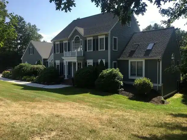 A large gray two-story house with vinyl siding replacement and white trim, features a gabled roof and is surrounded by shrubs and a lawn. A path leads to the front entrance in Brookline, MA, with another building visible in the background under a clear, sunny sky.