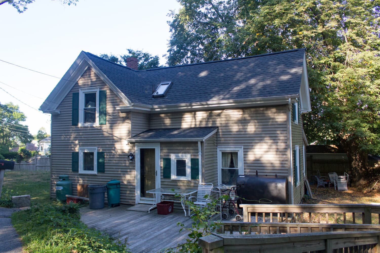 A two-story beige house with a dark roof and a small porch area. Theres outdoor furniture, a few garbage bins, and a bicycle nearby. The house is surrounded by greenery and trees. Shadows of trees can be seen on the houses exterior.