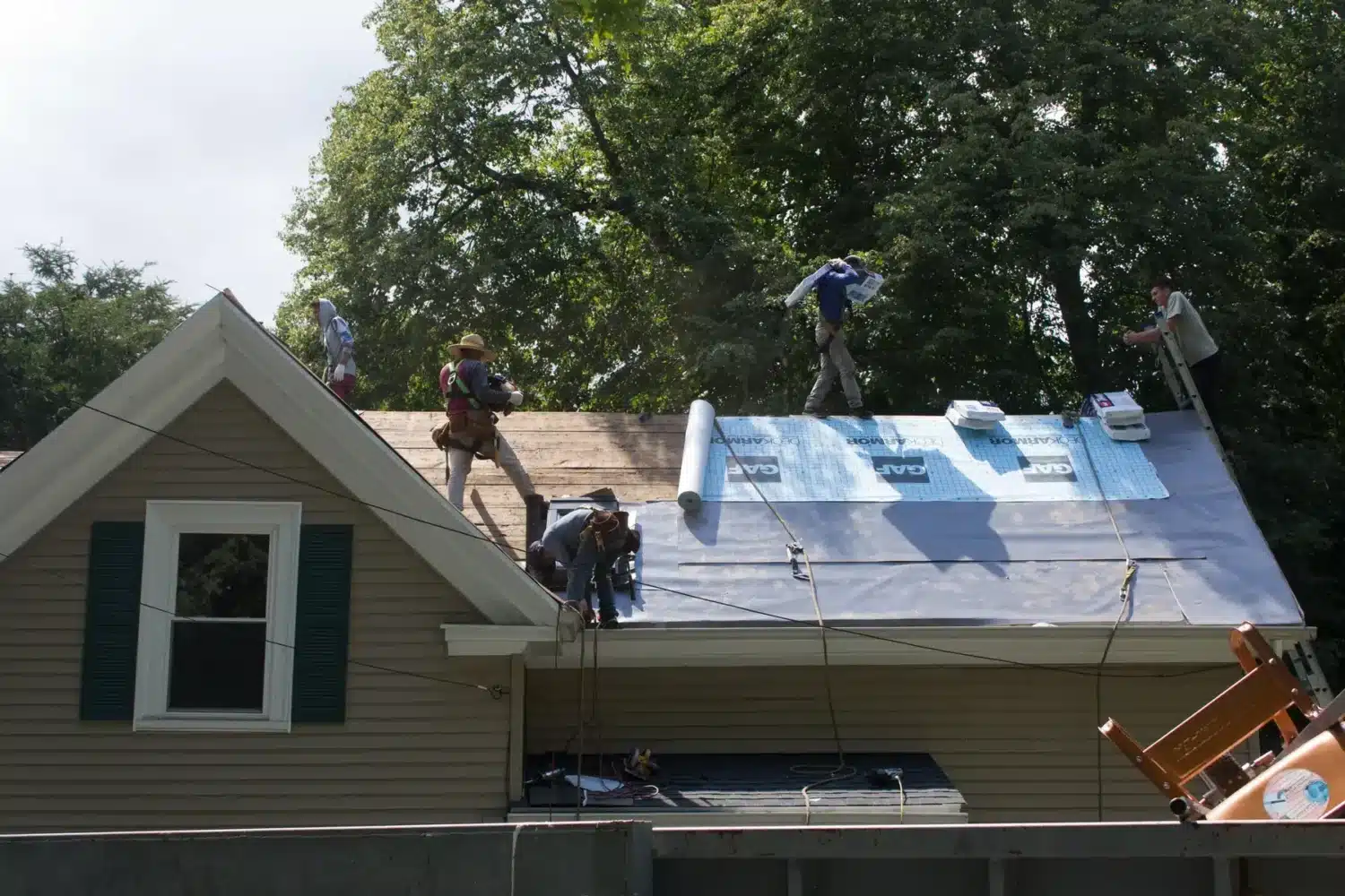 Four workers are installing new roofing material with leak barrier protection on a house roof, using tools and supplies scattered around them. The house is surrounded by trees, and the workers are wearing safety harnesses.