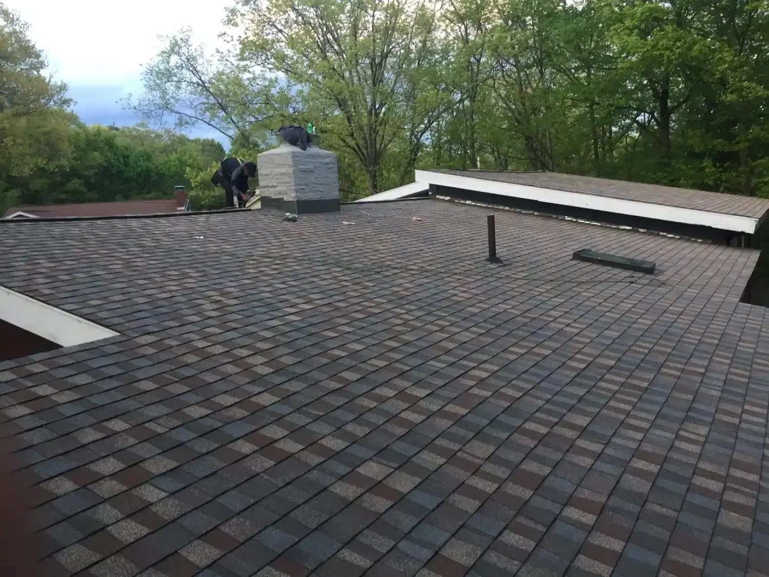A rooftop with new shingles in a pattern of dark gray and brown shades showcases quality craftsmanship. Two people, possibly highly-rated roofers, are working near a chimney in the background. Trees with lush green foliage surround the area under a partly cloudy sky.