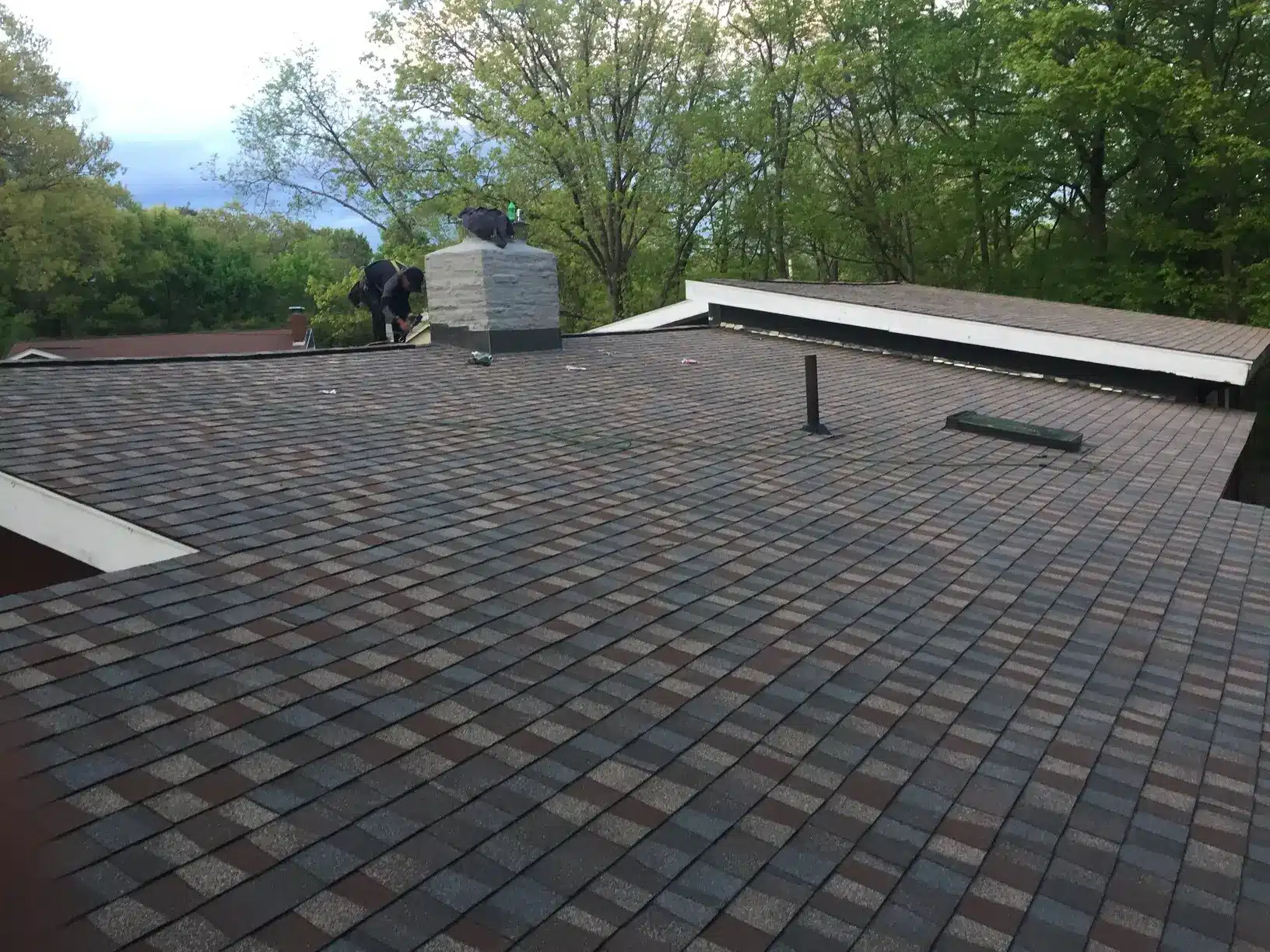 A roof covered with rectangular asphalt shingles in a staggered pattern; two people work near a chimney, while trees are visible in the background—an example often praised in roofer reviews.