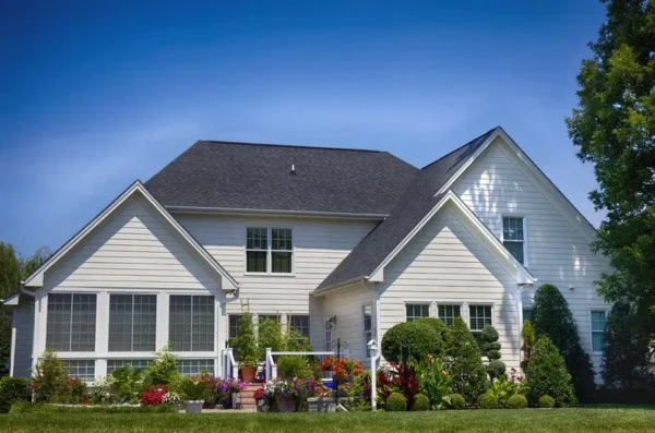 A charming white house with a dark roof, large windows perfect for enjoying the view, and a well-manicured front yard. The lush green grass, colorful flowers, and neatly trimmed shrubs thrive under a bright blue sky, making it an ideal scene to contemplate the best time of year to replace windows.