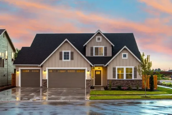 A suburban house with two stories, showcasing beige vinyl siding replacement in MA and a dark roof. It features two garages, a lit entrance, and a landscaped front yard. The sky in the background is a mix of pink and blue hues, suggesting sunset.