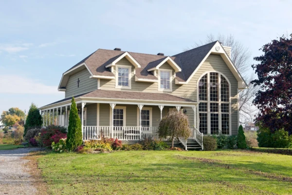 A charming two-story house with vinyl siding, large arched windows, and a wrap-around porch. Maintenance is a breeze in the well-kept yard filled with green grass, shrubs, and trees under a clear blue sky.