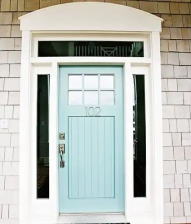 A light blue front door with the number 102 stands ready for winter, framed in white with slender vertical windows on each side and a row of small square windows at the top. The exterior wall is clad in light gray shingles, creating a welcoming yet sturdy entrance amidst the snowy season.