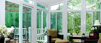 A bright sunroom with large windows and glass doors opening to a deck. This Three Season Porch features comfortable seating, a small table with a lamp, and lush greenery visible outside—perfect to add space and boost your home value.