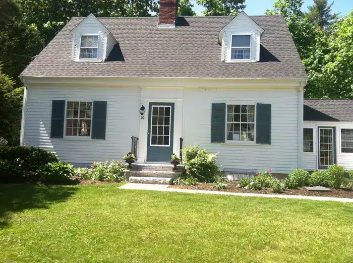 A white two-story house with a dark gray roof and dormer windows stands with timeless elegance. Its central front door, flanked by blue-shuttered windows, draws you in. A quaint porch fronts the neatly trimmed lawn from the 2014 Cost vs. Value Report era, where trees and shrubs add natural harmony.