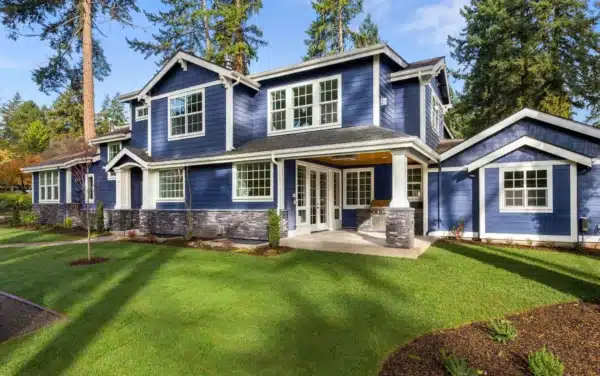 A two-story blue house with white trim sits surrounded by tall trees. The home features large windows ideal for a window replacement checklist. Stone accents on the lower exterior and a well-maintained green lawn flourish under a clear blue sky.