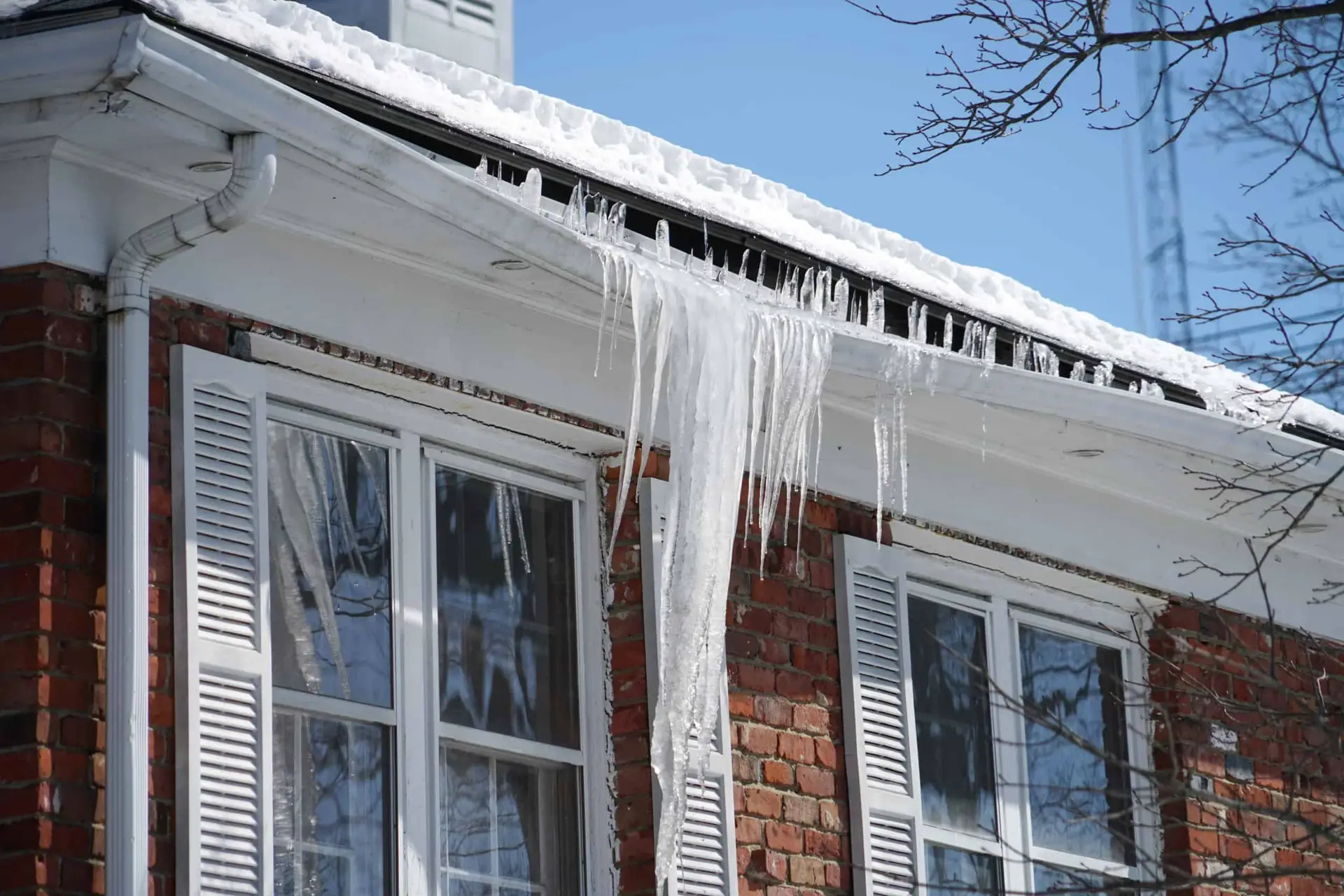 Large icicles hang from the roof edge of a red brick house with white shutters, above double windows—a classic view of roofing in winter. Snow blankets the roof, while bare tree branches frame a clear blue sky.