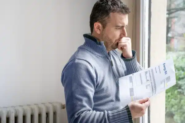 A man in a blue sweater stands by a winter window, looking worriedly at an energy bill in his hand. He rests his chin on his clenched fist, standing near a radiator with a view of green trees outside.