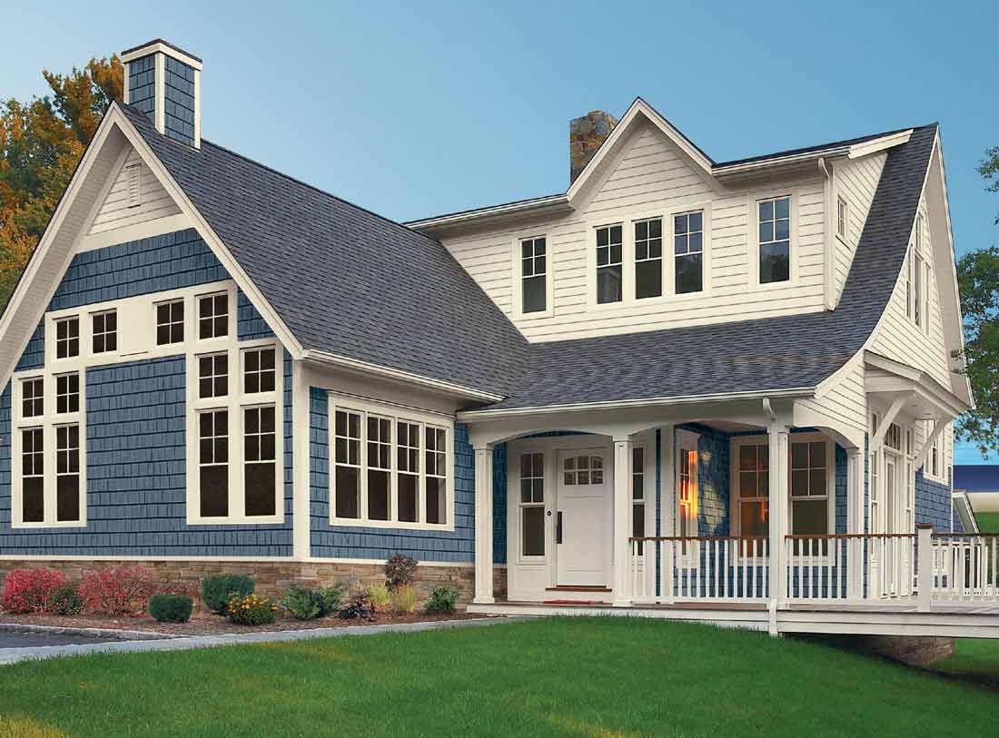 A two-story house with white siding and blue shingles features multiple windows, a stone foundation, and a welcoming porch. Amid the COVID-19 outbreak, enjoy peace of mind with our no-contact free estimate. The yard is green and well-maintained under a clear blue sky.