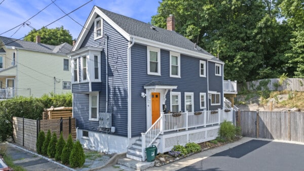 A charming two-story blue house with white trim and vinyl siding installation boasts a bright orange door. This Beverly, MA gem features multiple windows, a quaint front porch adorned with flower boxes, and a fenced yard rich in greenery. Nestled in a suburban area with trees and nearby homes.