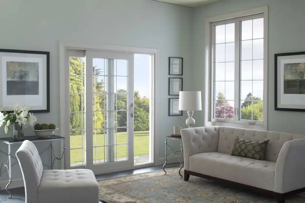 A bright living room with a beige sofa and chair, a floral rug, and a glass door leading to a green garden. Two large windows let in natural light. Framed art and a lamp on side tables adorn the space. White lilies are in a vase on a console table.