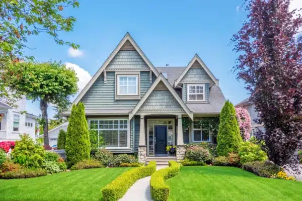 A well-maintained suburban house with a gray facade, steep gable roofs, and white trim awaits your spring home improvement touch. The front yard features manicured hedges, lush green grass, and various shrubs and trees against a clear blue sky.