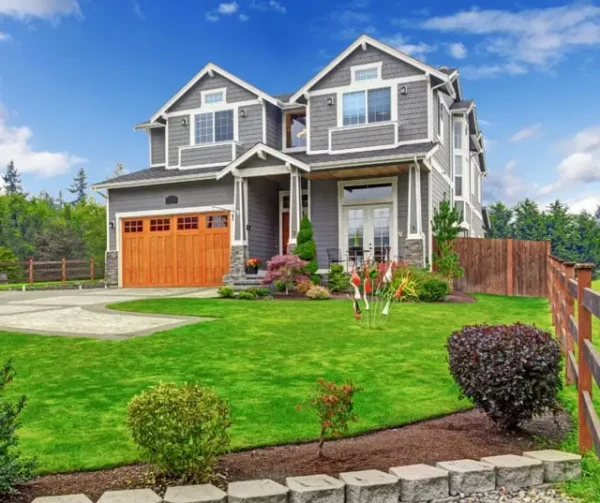 A two-story, modern gray house with large windows and a wooden garage door. The neatly landscaped front yard showcases the benefits of Spring, with a lush green lawn, shrubs, and colorful flowers. A wooden fence runs along the right side under a clear blue sky.