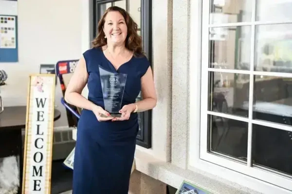 A woman with long brown hair in a navy blue dress stands smiling, holding her Business Person of the Year award. She is in an office space near a window, with a WELCOME sign and various office items visible in the background, captured beautifully by Salem News.