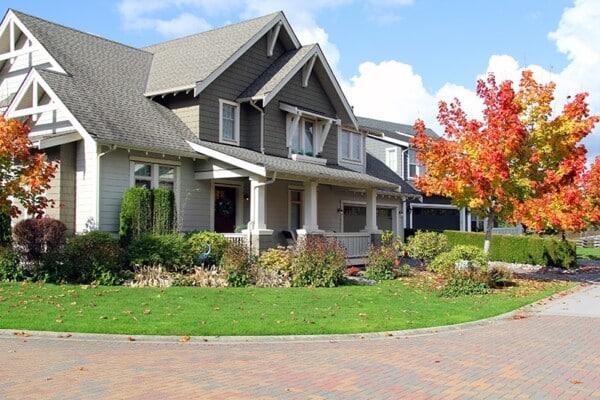 A charming suburban house with a grey exterior, white trim, and a covered porch showcases impeccable fall maintenance. A well-maintained lawn and vibrant red-orange autumn tree enhance the scene. The driveway is paved with bricks, and the sky is partly cloudy.