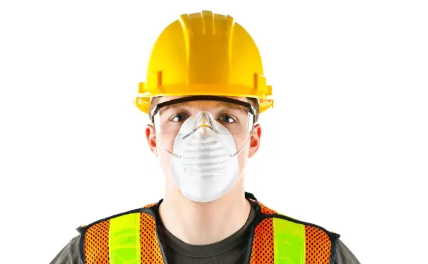 A person wearing a yellow construction helmet, safety goggles, an orange high-visibility vest, and a white dust mask stands against a white backdrop, ensuring every detail aligns with the strict standards similar to those mandated by Exteriors Ordinance for public safety and compliance.