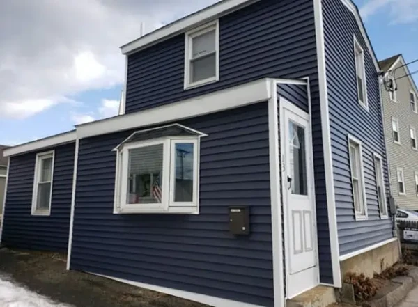 A blue two-story house with white trim and a small overhang above the front door proudly showcases its bay window. Vinyl siding complements the exterior, where a mailbox is attached. The sky is partly cloudy, lending just the right backdrop to this charming installation.