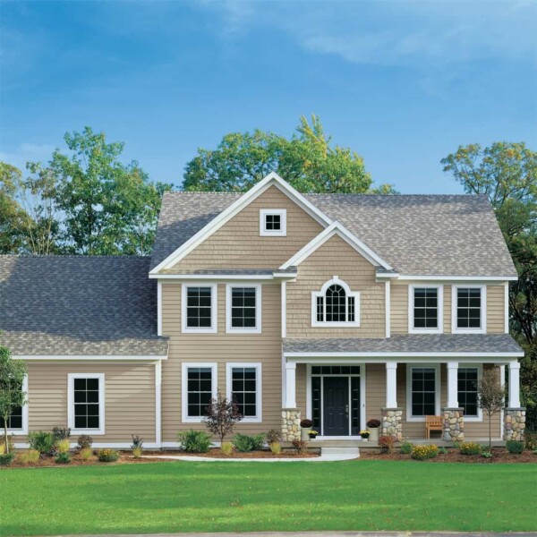 A charming New England home with beige vinyl siding, white trim, and a gray roof. It features a symmetrical design with multiple windows, a central entrance adorned with columns, and a cozy porch. A lush green lawn and trees stretch across the background under a clear blue sky.