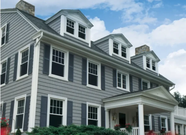 A large, three-story gray house with white trim, vinyl cedar shakes, dark shutters, and dormer windows on the roof; there is a covered front porch and some greenery in the foreground, set against a blue sky with clouds.