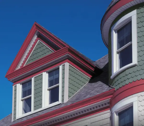 A close-up of a Victorian-style house with green fish scale shingles, white trim, red accents, and replacement windows, featuring a gabled roof and a rounded turret under a clear blue sky.