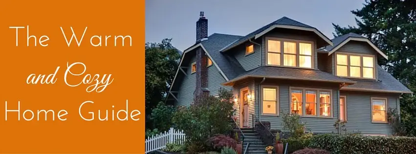 A two-story cozy home with warm glowing windows at dusk, surrounded by greenery and a white picket fence. Text on an orange background reads: The Warm and Cozy Home Guide by Beverly Patch.