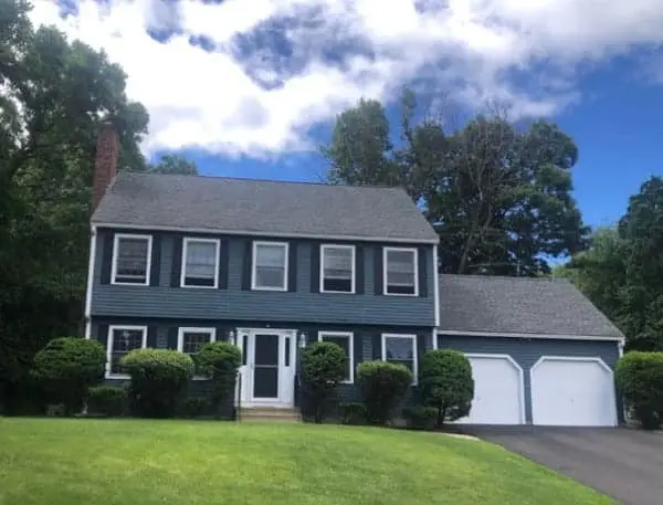 A two-story blue house with white trim, featuring a two-car garage, embodies summer home improvement ideas. Surrounded by lush green bushes and trees under a bright blue sky with clouds, it boasts a well-kept lawn that completes its inviting charm.