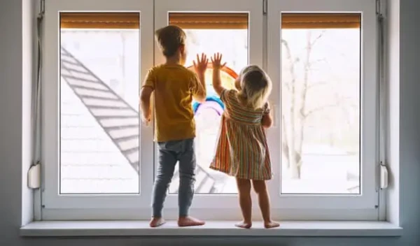 Two young children stand on a decorated windowsill, gazing outside in wonder. The child on the left sports a yellow shirt and jeans, while the one on the right dons a striped dress. Their hands press against the glass, serving as a gentle reminder of National Window Safety Week.