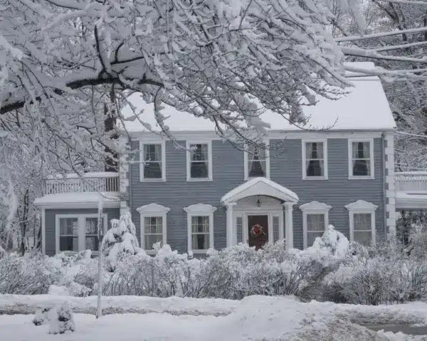 A two-story blue-gray house with white trim is surrounded by snow-covered trees and bushes. Snow blankets the ground and roof, showcasing a peaceful scene that highlights the homes exterior in cold weather.