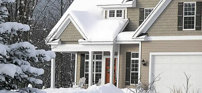 A house with a snow-covered roof, surrounded by snow-covered trees and yard. Two white chairs sit on the front porch near the entrance, next to a white garage door—perfect for discussing winter home safety tips.