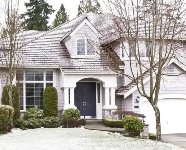 A light gray suburban house boasts a shingled roof, large windows, and a dark blue front door. The home improvement contractors touch is evident in the trimmed bushes and trees. A leafless tree stands prominently in the foreground on a crisp winter day.