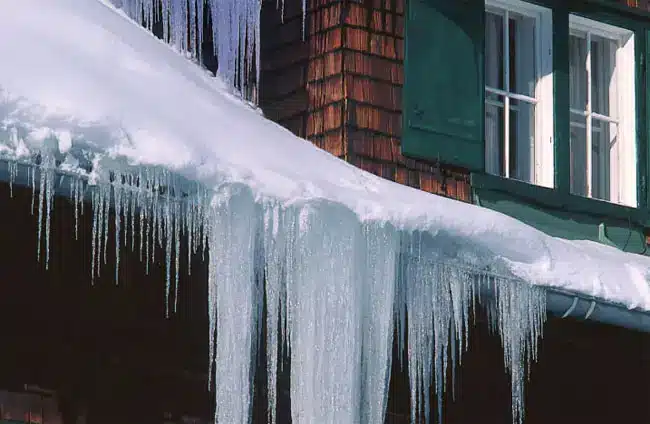 Large icicles hang from the snow-covered edge of a roof on a building with wooden shingles and green window shutters, suggesting cold winter weather and the potential for ice dams to form.
