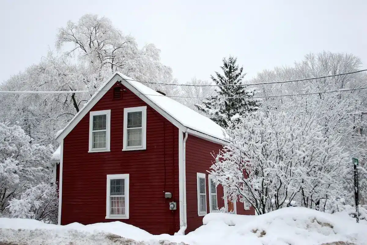 A red wooden house is surrounded by a snowy landscape, offering a picturesque winter scene as trees and bushes are blanketed in snow. The sky is overcast, enhancing the chilly ambiance—a perfect reminder to winterize your home against the cold.