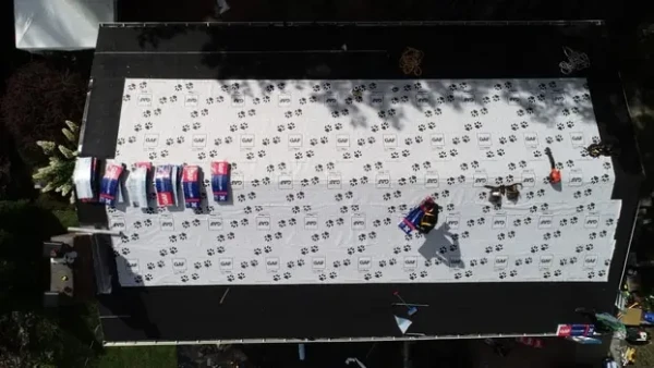 Aerial view of a roof under construction in Woburn, MA. The roof is covered with white underlayment, marked with dark logos for superior protection from the elements. Several bundles of roofing material are stacked on the left side, while workers cast shadows on the right.