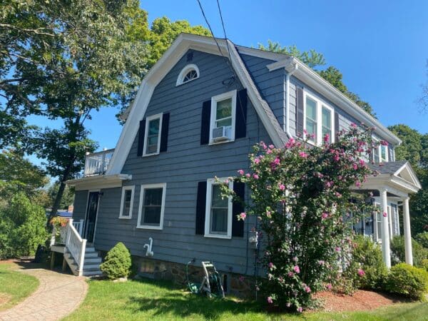 A two-story house with gray siding and white trim, featuring a gambrel roof and dark shutters. A flowering bush with pink flowers grows near the side. There are trees and a green lawn with a walkway leading to steps at the front.