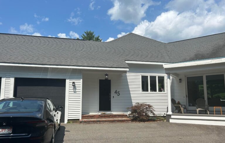A white house with black shutters and roof, the number 45 beside the door, a black car in the driveway, and a covered porch with two chairs on the right—an inviting scene perfect for a Roofing Contractor in Bedford MA. The sky is partly cloudy.