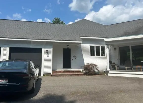 A single-story white house with a gray roof, installed by a trusted Roofing Company in Chestnut Hill, a black car parked in front of the garage, the number 45 by the entrance, and a covered porch with two chairs under a partly cloudy sky.
