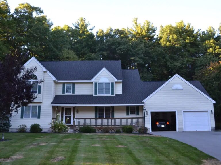 Two-story suburban house with cream-colored siding, black roof, front porch, and attached two-car garage. The lawn is partially mowed with trees behind. Quality roofing in Chestnut Hill ensures a sturdy black roof. One car is parked inside the garage.